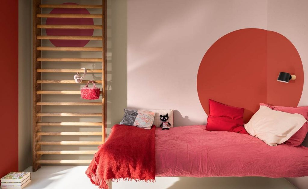 Camera da letto con parete rosa e rossa, scala di legno appoggiata sul muro. Letto con lenzuola e cucini rossi.