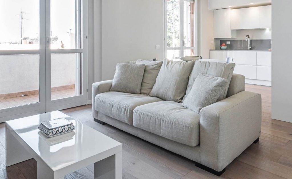 Divano due posti grigio chiaro con tavolino in legno bianco lucido. Pavimento con rivestimento in parquet effetto legno chiaro.